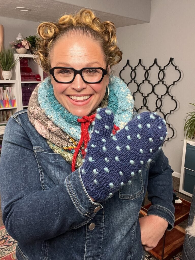 A smiling person with curly hair, glasses, and a denim jacket shows off knit mittens made from a free knitting pattern. They wear a multi-colored knit scarf and stand in a cozy, decorated room inspired by the Andes. -Marly Bird