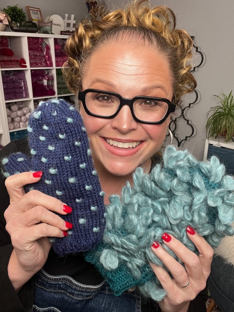 A smiling woman with curly hair and glasses holds up two colorful Andes Thrummed Knit Mittens in a cozy room, surrounded by yarn and plants—perfect inspiration for your First Fall 2025 Make-Along with this free knitting pattern. -Marly Bird
