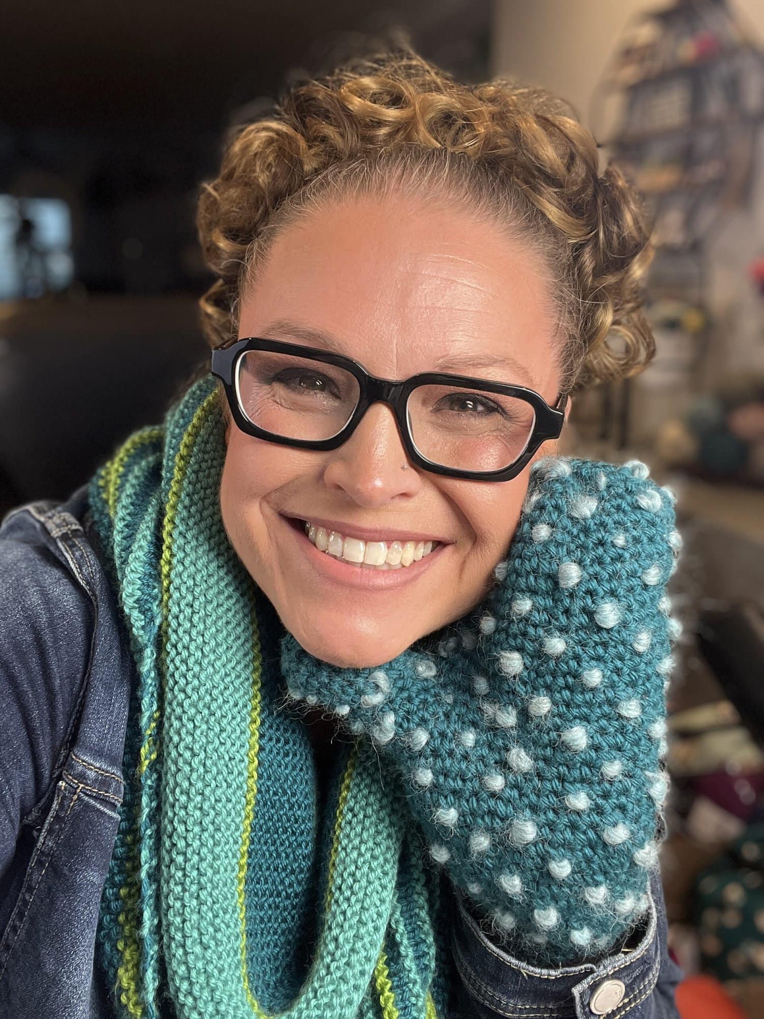 A smiling person with curly hair and glasses wears a blue and green knit scarf and a textured blue knit mitten, posing for a cozy close-up selfie indoors. -Marly Bird