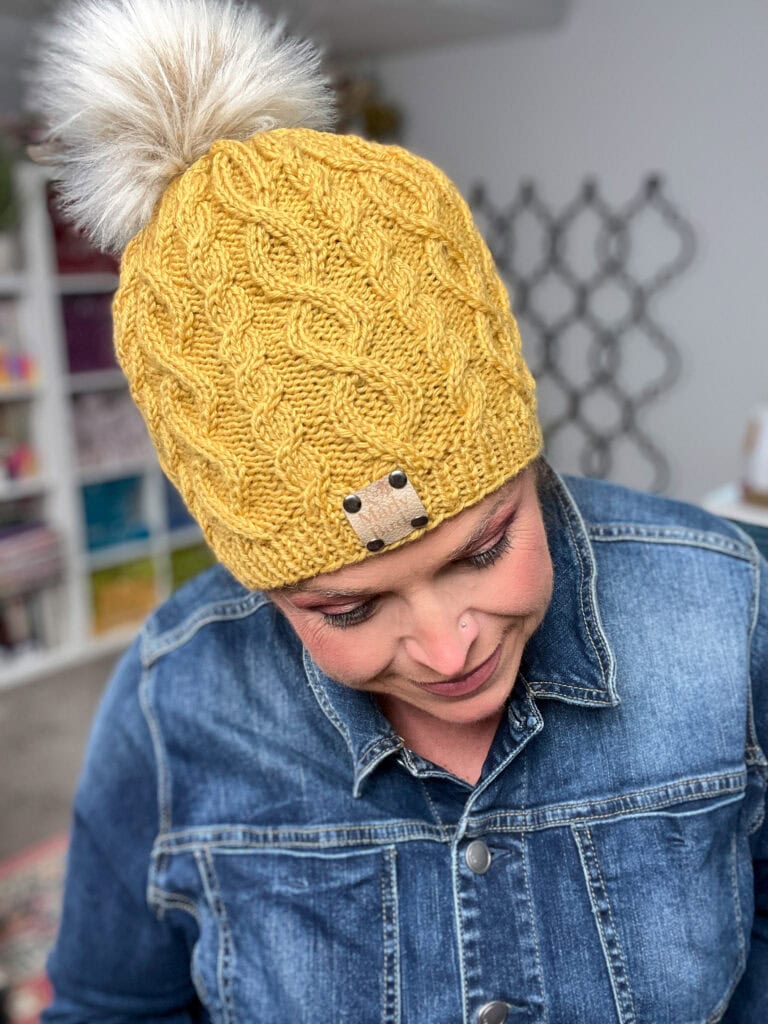 A person in a blue denim jacket looks down, wearing the Keystone Cabled Knit Hat—a yellow knit hat with a cable pattern and large faux fur pom-pom—perfect for Turkey Trot 2025. Shelves and soft, homey decor are visible in the background. -Marly Bird