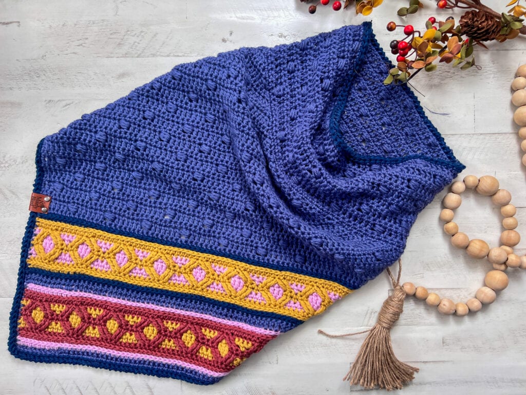 A crocheted shawl with a textured blue body and multicolored patterned stripes in yellow, pink, and maroon at the edge, displayed on a light surface with wooden beads and decorative branches; inspired by Ridgeway Crochet Cowl gauge. -Marly Bird