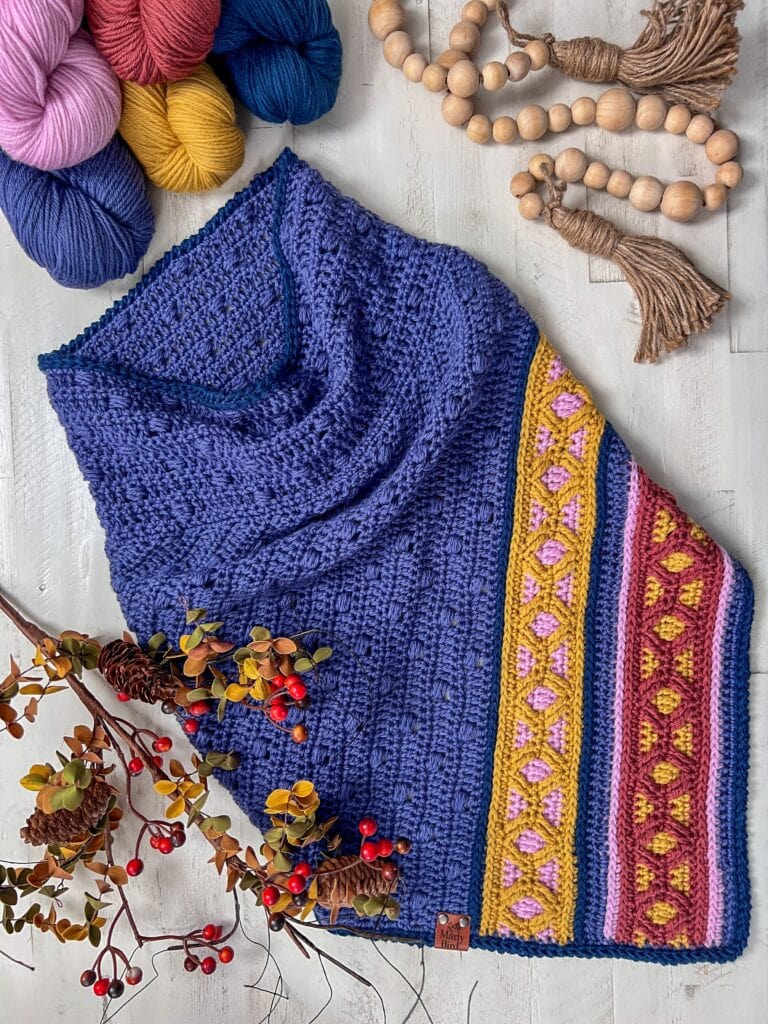 A Ridgeway Crochet Cowl in deep blue with colorful pink, yellow, and green patterned stripes lies on a white surface, surrounded by yarn balls, wooden beads with tassels, and sprigs of red berries. Perfect for your next crochet pattern project!. -Marly Bird