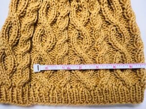 A close-up of a yellow knitted fabric with a cable pattern, featuring a pink and white measuring tape laid horizontally across the fabric, illustrating a gauge swatch guide for the Keystone Cabled Knit Hat at about 11 inches. -Marly Bird