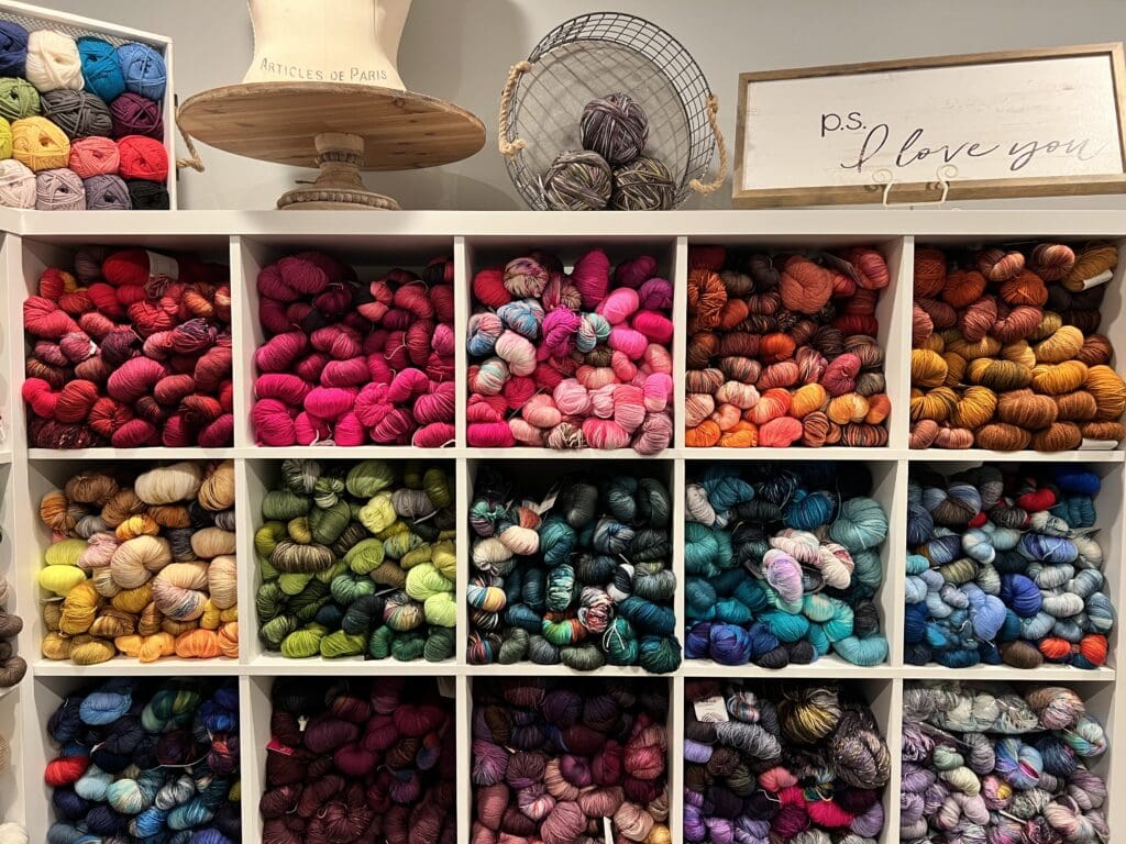 A shelf brimming with vibrant yarn skeins, neatly organized by shade from pinks to oranges, greens, blues, and neutrals. A crochet hat form adds charm, while a wire basket brims with yarn. A sign sweetly declares, "P.S. I love you," hinting at a crafty spring cleaning. -Marly Bird