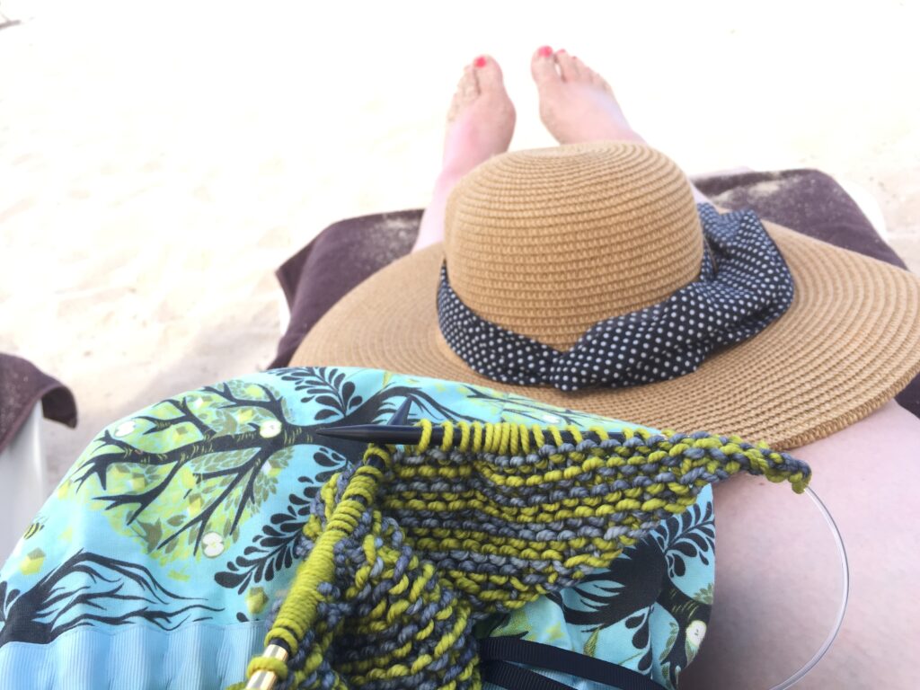 knitting needles in a work in progress shawl sitting on the lap of Marly Bird next to a sun hat - small knitting projects are great for travel - yes knitting needles can go on a plane - Marly Bird