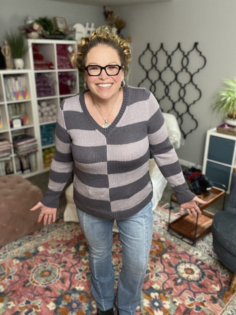 A smiling woman with curly hair and glasses stands indoors on a colorful rug, wearing jeans and her favorite knit sweater pattern. Shelves with yarn and books, plants, and home decor decorate the cozy background. -Marly Bird
