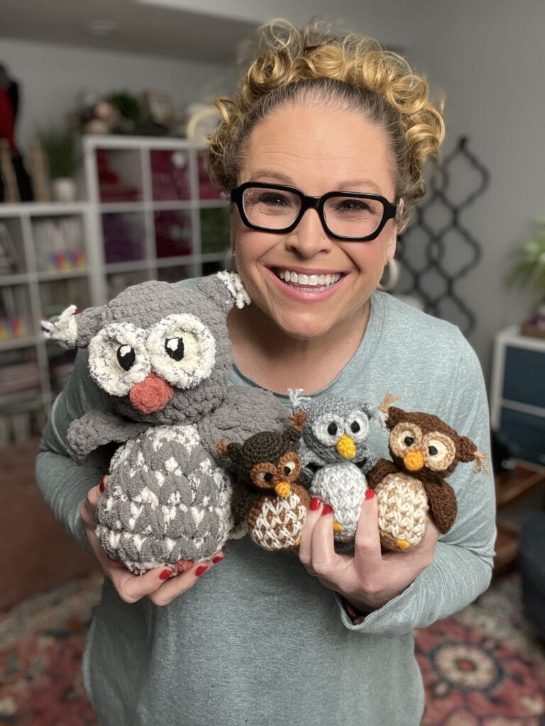 A smiling woman with glasses holds five crocheted owl toys of varying sizes and colors in her hands, standing in a cozy, well-lit room with shelves and plants—perfect inspiration for those seeking free knitting and crochet patterns. -Marly Bird