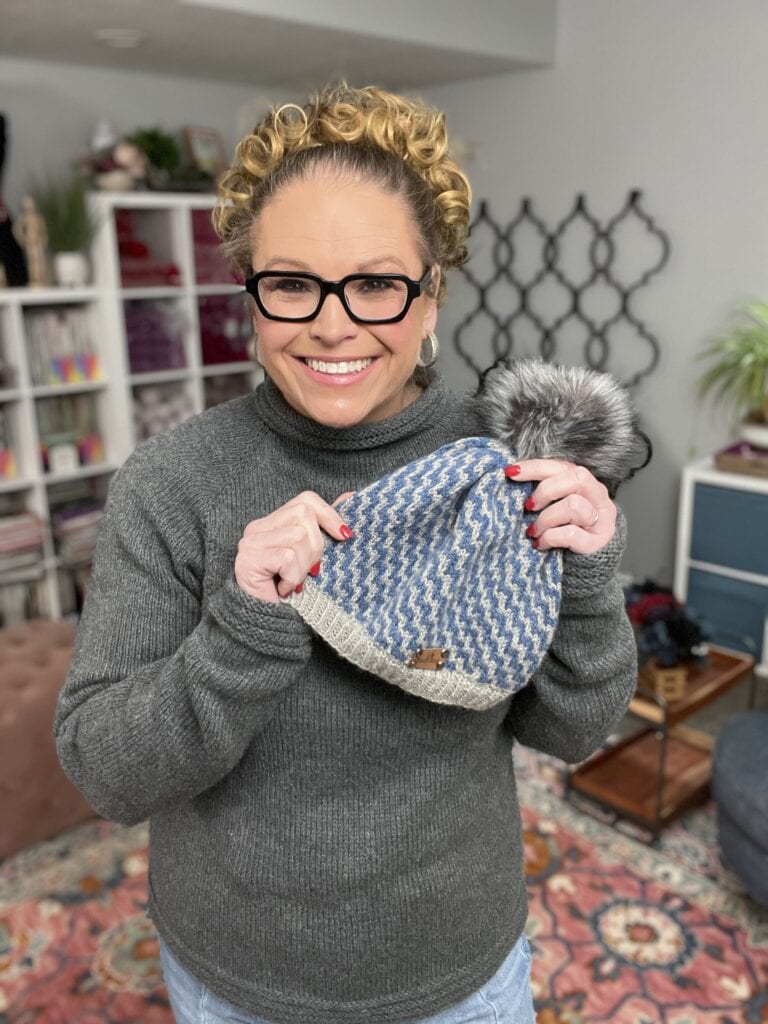 A smiling person with curly hair, glasses, and a gray sweater holds a blue and gray knit hat with a faux fur pom-pom—created using free knitting and crochet patterns. They're in a cozy, well-lit room with bookshelves and colorful decor. -Marly Bird