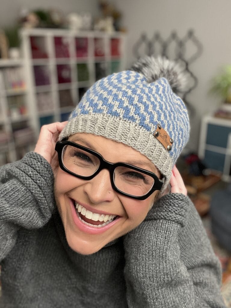 A smiling person wearing black glasses, a blue and gray knit beanie with a pom-pom, possibly made from free knitting and crochet patterns, and a gray sweater holds the sides of the hat. The background shows a room with shelves and various items. -Marly Bird