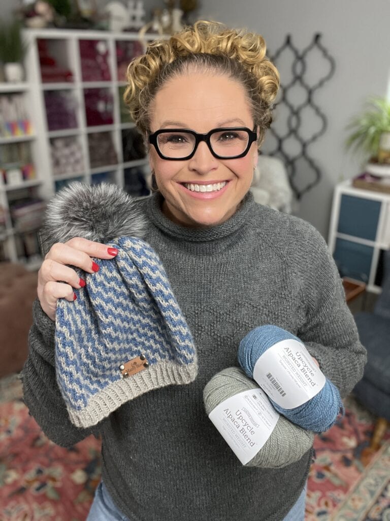 A smiling woman with curly hair and glasses holds a blue and gray knit hat with a pom-pom in one hand and two balls of alpaca yarn in the other, surrounded by shelves and plants—perfect inspiration for free knitting and crochet patterns. -Marly Bird