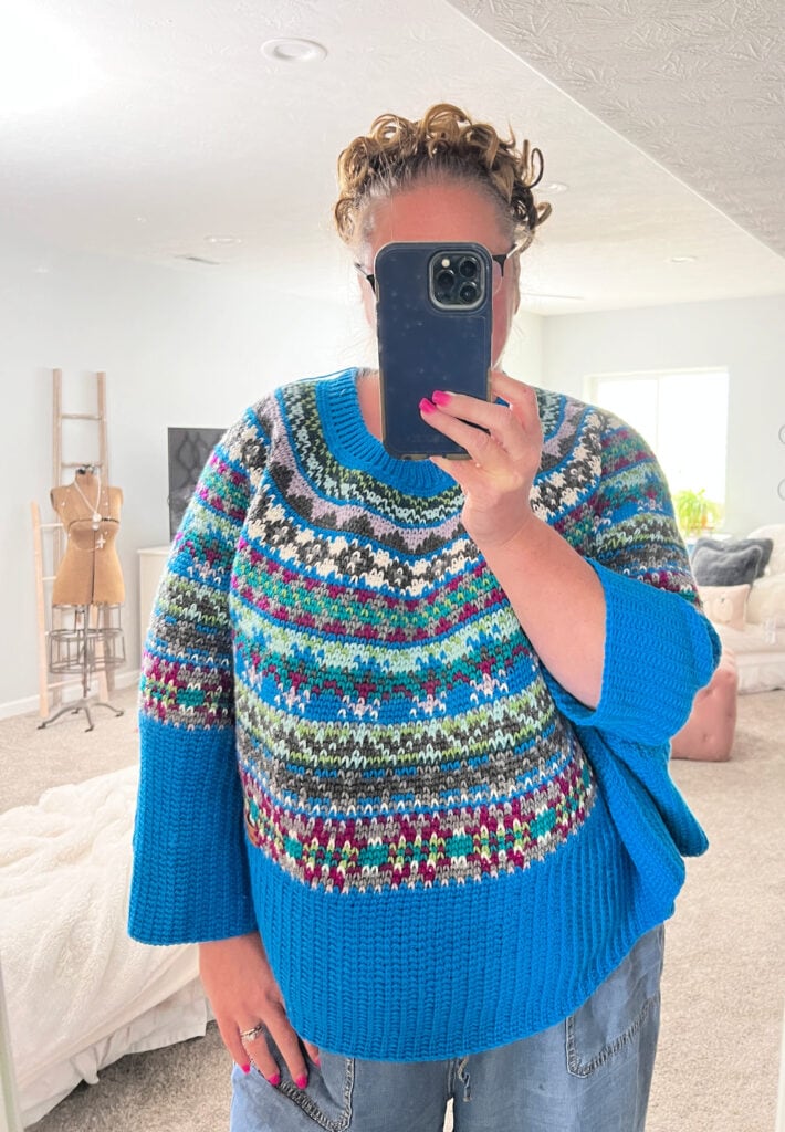 A person with curly hair takes a mirror selfie in a colorful Prism Make-Along knit sweater of blue, green, white, and burgundy. Holding a phone with a blue case, they stand in front of a bright, tidy bedroom. -Marly Bird