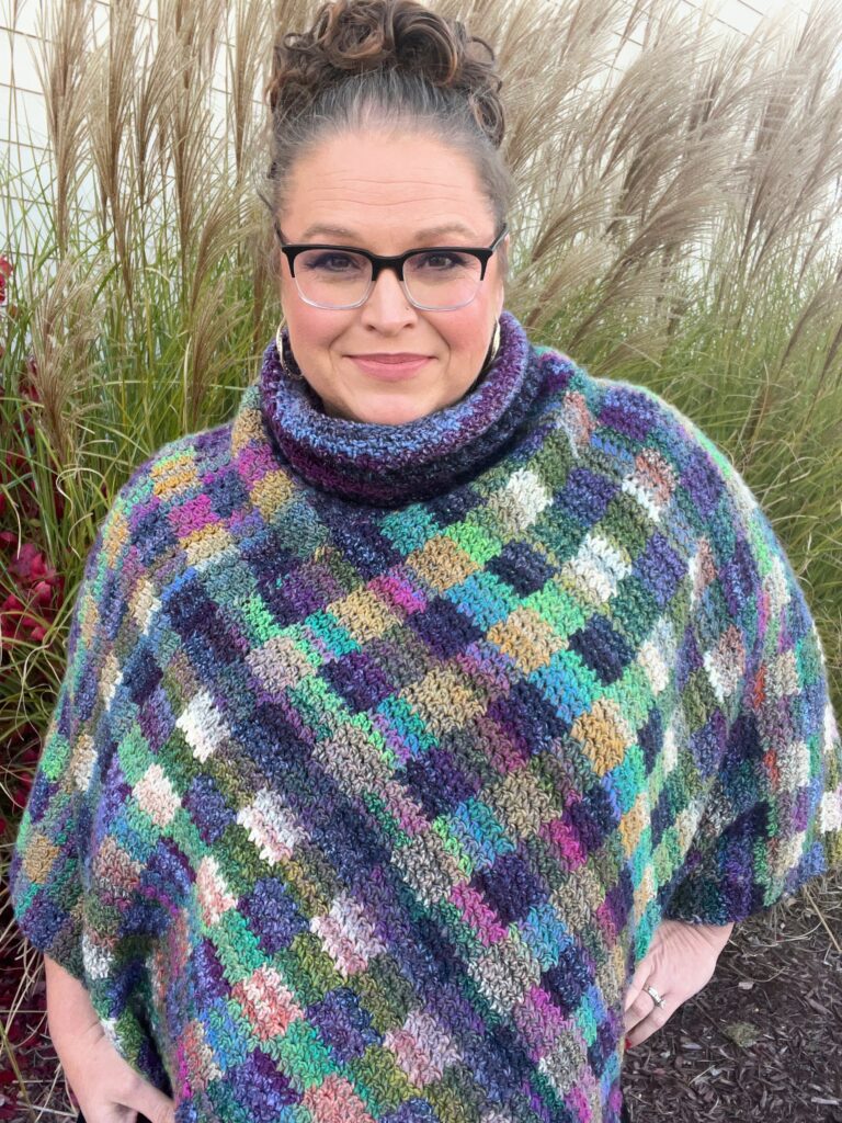 
A woman stands outdoors, smiling at the camera, wearing a crocheted poncho featuring a colorful gingham plaid pattern. The poncho has a cowl neck and a variety of hues including purple, green, and blue, creating a cozy and vibrant look. She's accessorized with glasses and has her hair styled up, with a backdrop of tall grasses. Marly Bird Check Me Out Crochet Poncho




