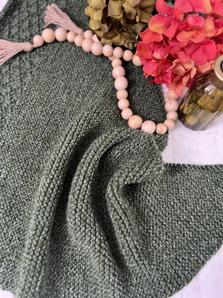 A textured green woven fabric lies on the surface, alongside a string of wooden beads and pink flowers. A small glass bottle adds a decorative touch to this tranquil arrangement, reminiscent of the Peak Serenity often found in a cozy knit shawl. -Marly Bird