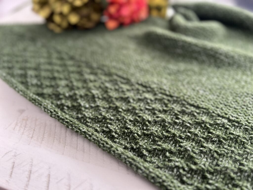 Close-up of a textured green fabric with a patterned knit design reminiscent of a Serenity Knit Shawl. In the blurred background, colorful flowers, including orange and yellow blooms, add to the cozy, handmade feel of peak serenity. -Marly Bird