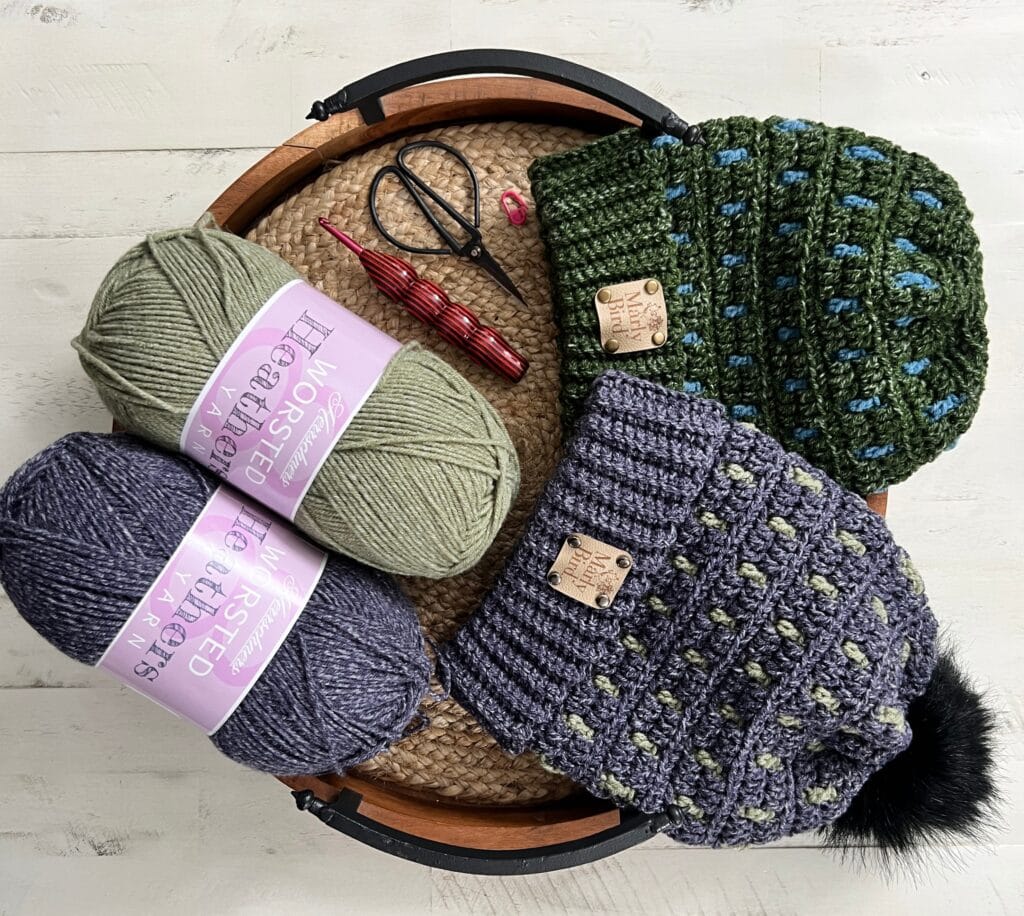 A round basket holds a green crocheted hat with blue accents and a purple hat with a black pom-pom. Perfect for 2025 game day, two skeins of yarn, one green and one purple, along with crochet hooks and stitch markers rest beside the hats on a light wooden surface. -Marly Bird