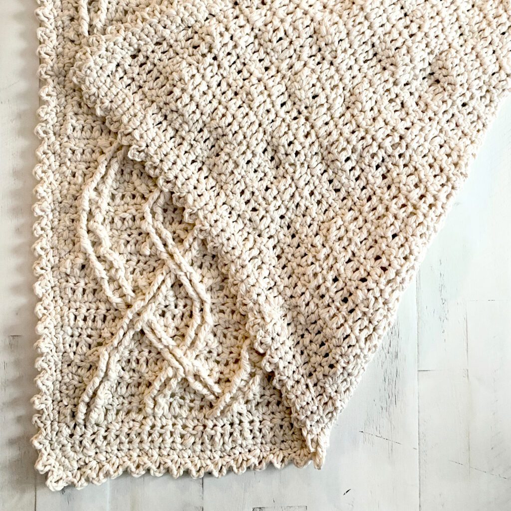 A cream-colored crocheted cabled blanket with a textured pattern and picot edging, partly spread out on a white wooden surface to show the cabled right side and smooth wrong side of fabric. Inishmore Crochet Cabled Blanket by Marly Bird.