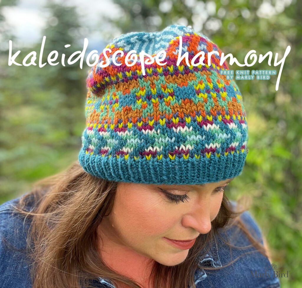 The image shows a person wearing a hand-knit hat with intricate colorwork. The hat, named "Kaleidoscope Harmony," features a vibrant mix of colors creating a striking pattern. The text on the image reads "kaleidoscope harmony free knit pattern by Marly Bird," indicating that this is a design by Marly Bird and that the pattern is available for free. The colorful design of the hat is reminiscent of a kaleidoscope with its symmetrical, repeating patterns and a harmonious blend of colors.