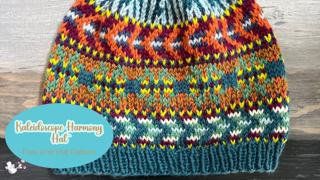 The image showcases a beautifully knitted hat laid flat on a wooden surface, providing a clear view of the intricate colorwork pattern. The pattern features an array of vibrant colors forming a complex, kaleidoscopic design. The lower part of the hat has a ribbed band in a teal color, which provides a nice contrast to the multicolored body of the hat. Overlaying the image is a stylized text that reads "Kaleidoscope Harmony Hat," followed by "Free Knit Hat Pattern," indicating that the pattern for this hat is available for free. The design elements and color scheme of the hat are typical of Marly Bird's distinctive and colorful style in knitwear.