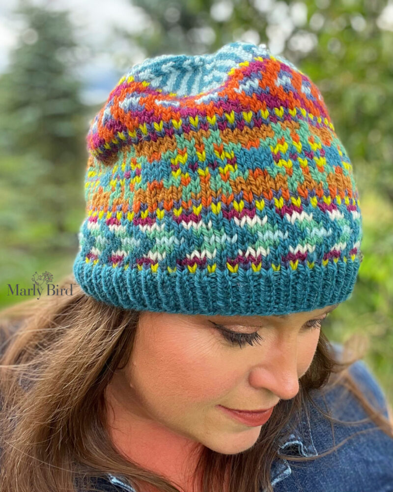 A woman with her head tilted down, showcasing a colorful colorwork knit hat. The hat features vibrant patterns in shades of blue, orange, and green. -Marly Bird