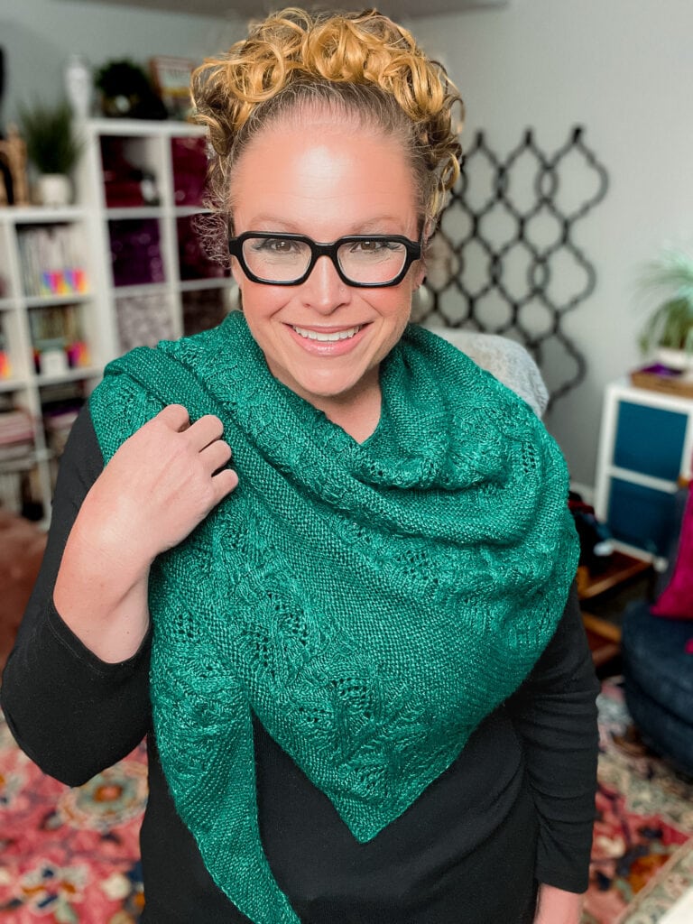 Marly Bird models a textured green knit boomerang shawl with bold stitch definition, worn over a black top in a cozy room. 