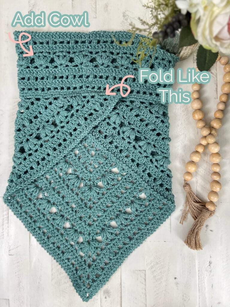 A textured, teal crocheted triangle is laid flat. Arrows and the text "Kembleford Crochet Cowl" are at the top, with "Fold Like This" in the center. A wooden bead and tassel adornment rests beside it, surrounded by delicate white flowers. -Marly Bird