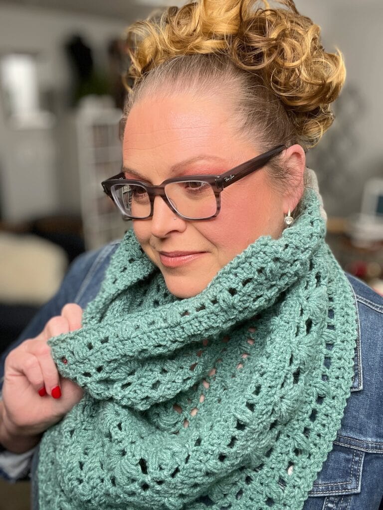 A person with curly hair wearing glasses and a denim jacket dons a Kembleford Crochet Cowl. Indoors, they hold up the textured green scarf with one hand, maintaining a calm expression. -Marly Bird