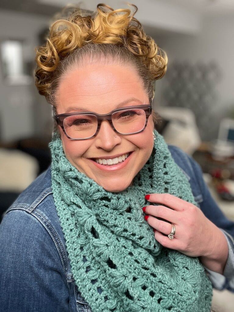 A person with curly hair and glasses smiles warmly at the camera, wearing a denim jacket and a crocheted green cowl. Red nail polish brightens their fingernails, while the background is softly blurred in the charming style of Kembleford photography. -Marly Bird