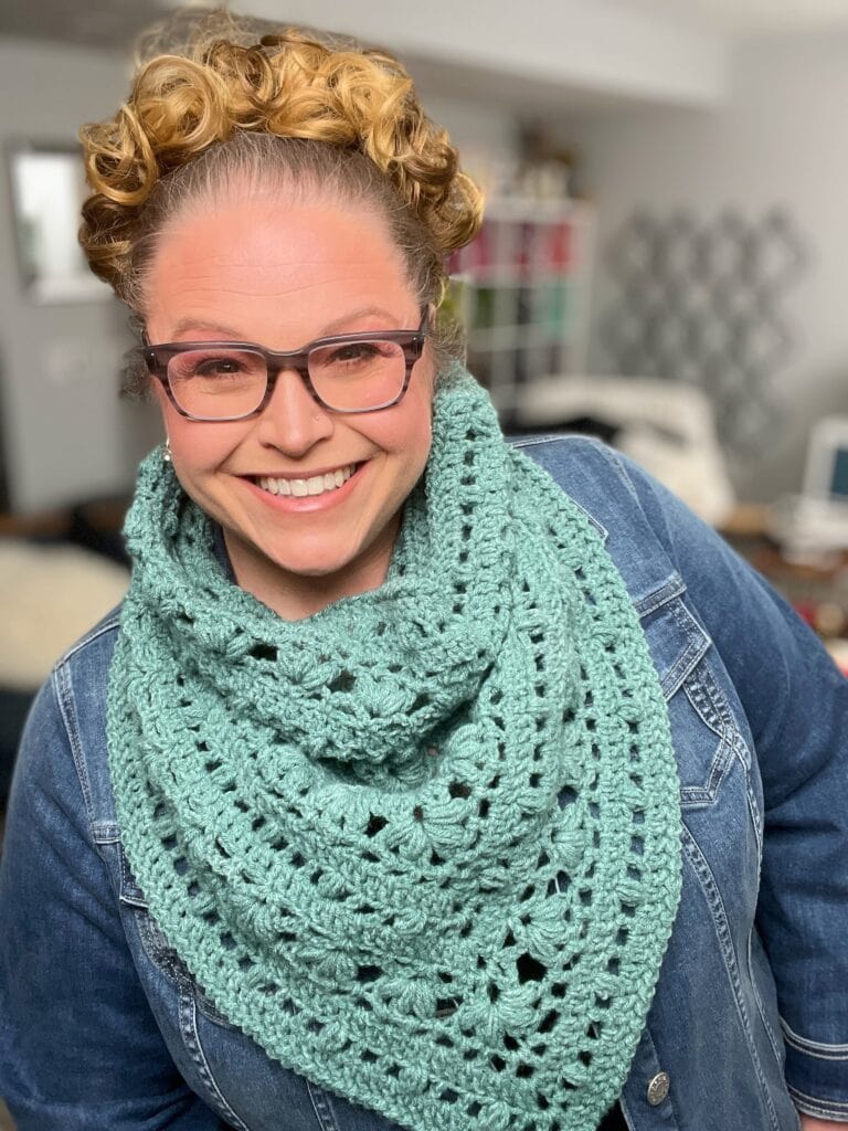 A person with curly hair and glasses is smiling. They are wearing a blue denim jacket and a large, textured mint green crochet scarf. The background is blurred, with some shelves and geometric patterns visible. -Marly Bird