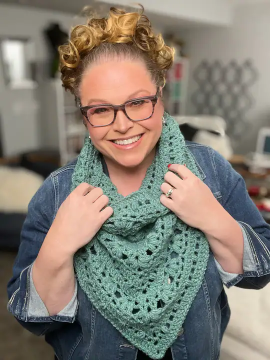 A smiling person with curly hair and glasses wears a blue denim jacket and holds a light green crocheted scarf—perfect for those seeking free knitting and crochet patterns—around their neck indoors. -Marly Bird