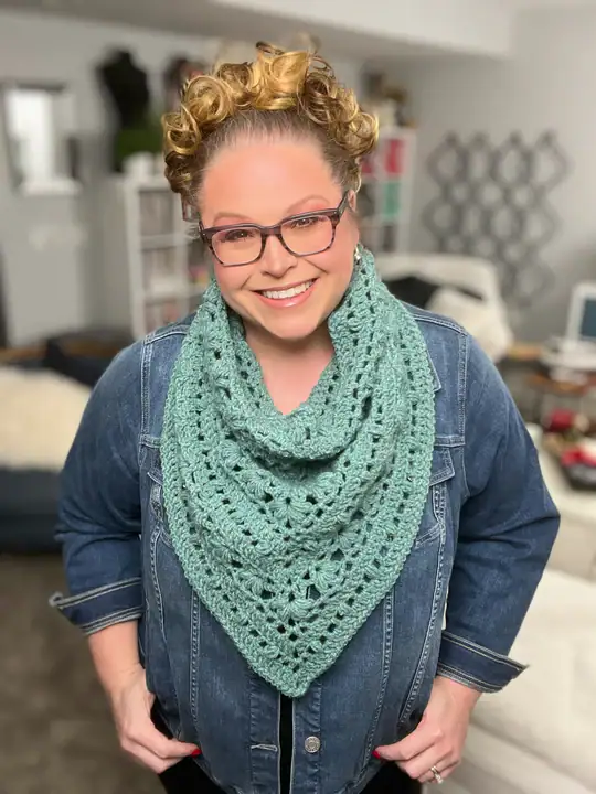 A smiling woman with curly hair and glasses wears a denim jacket and a light green crocheted scarf, standing in a cozy living room. Her stylish look is perfect inspiration for those searching for free knitting and crochet patterns. -Marly Bird