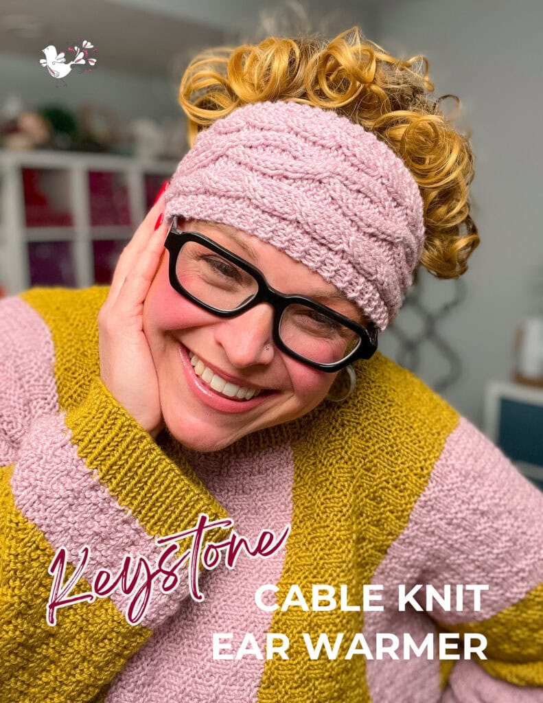 A smiling person with curly hair, black glasses, and a pink cable knit ear warmer poses with their hand on their face. They wear a pink and yellow sweater from the Jolly Holiday collection. Text reads: "Keystone Cable Knit Ear Warmer. -Marly Bird