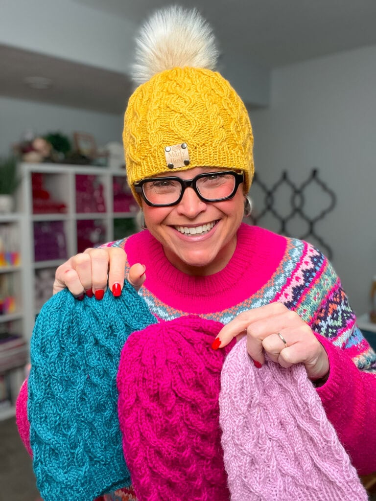 A smiling person wearing a yellow knit hat, glasses, and a colorful sweater holds up three Keystone Cabled Knit Hats in teal, pink, and light purple. Shelves with yarn are visible in the background for Day 5 of Turkey Trot 2025. -Marly Bird