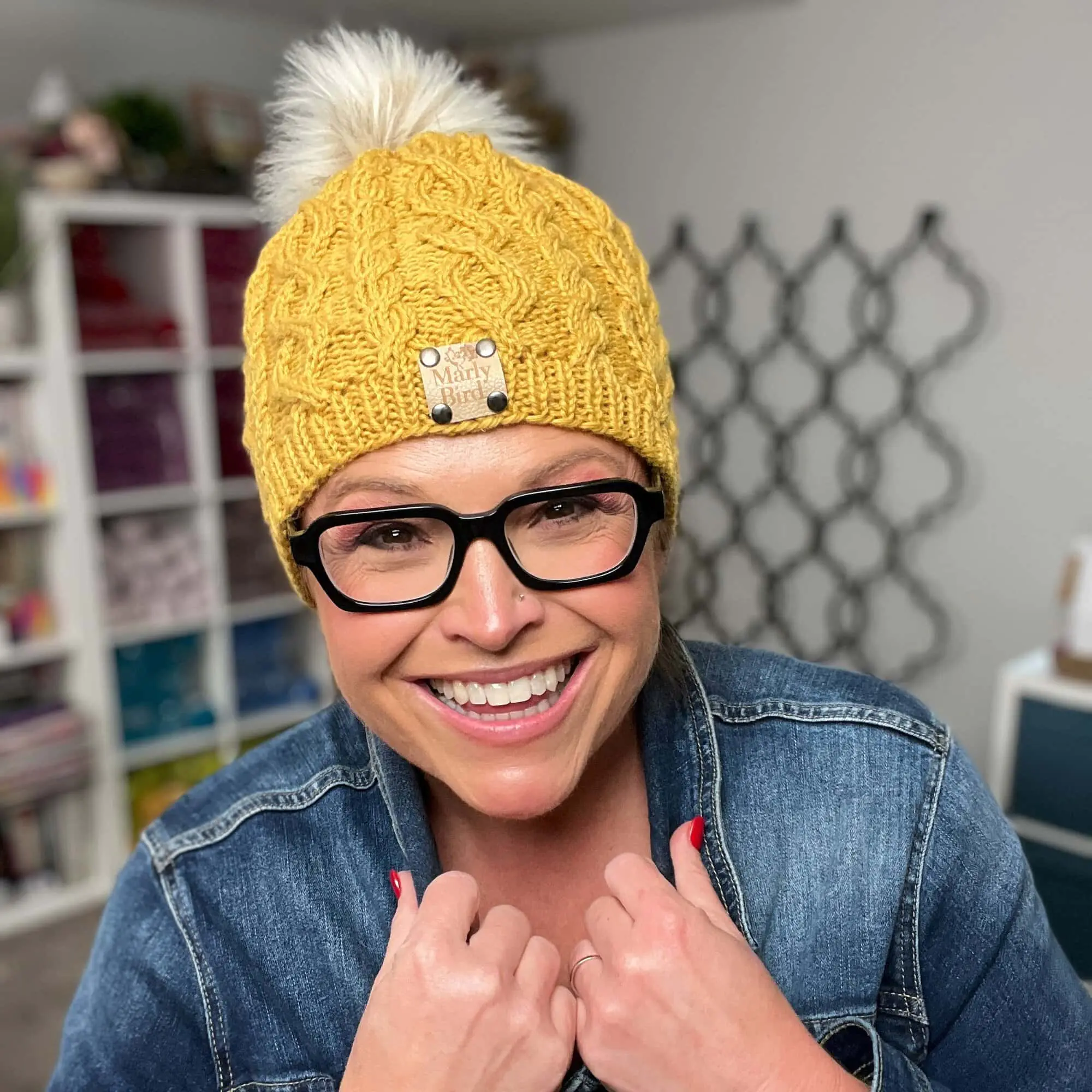 A person models a yellow, hand-knit cable hat with a white pom-pom and denim jacket; textured stitches are visible.