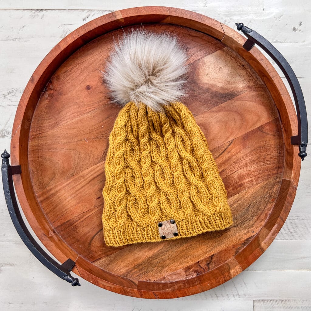 A mustard yellow knitted beanie with a thick cable pattern and a large, fluffy pom-pom on top, placed inside a round wooden tray with black metal handles—perfect for your Marly Bird Turkey Trot Make-Along 2025 project. -Marly Bird