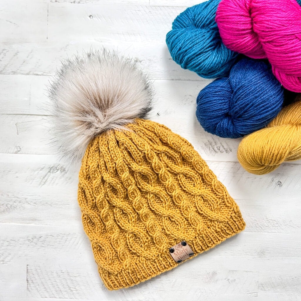 A yellow knit hat with a faux fur pom-pom is displayed on a white wooden surface, next to skeins of blue, fuchsia, and yellow yarn—perfect for the Marly Bird Turkey Trot Make-Along 2025. -Marly Bird