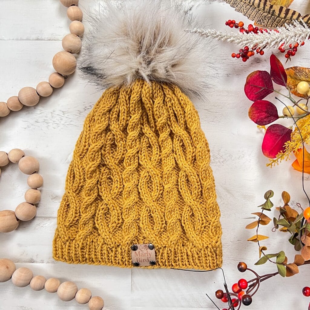 Mustard yellow cable knit hat with faux fur pom-pom, displayed on white wood and accented by autumn beads, berries, and leaves.
