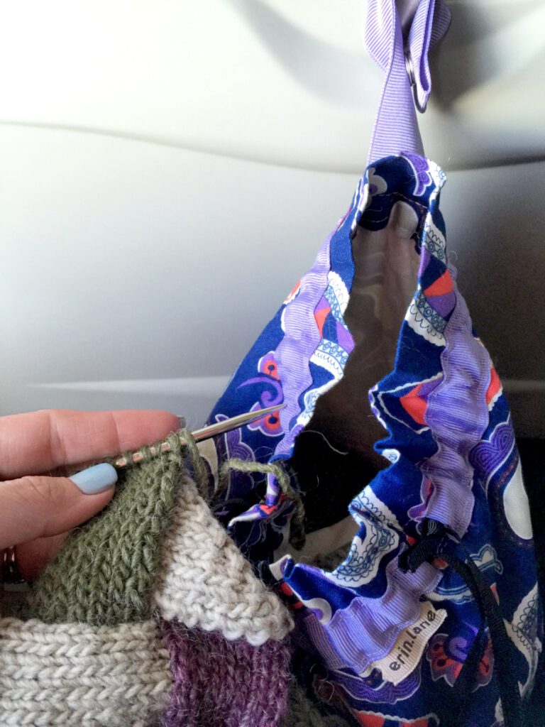 Knitting needles on a plane and an Erin Lane Bags Twofer bag hanging from the tray table clip - knitting needles with entrelac knitting - Marly Bird