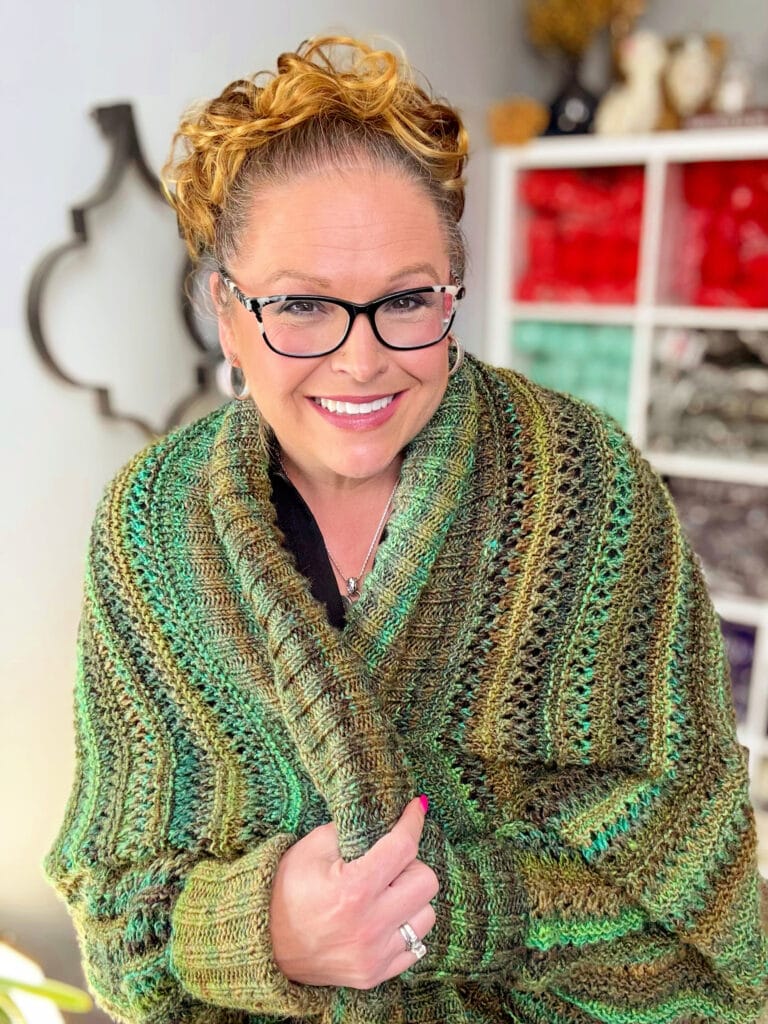 Marly Bird modeling the Know Your Worth Knit Cocoon Cardigan in the Lace Bands stitch pattern, the cocoon-cardigan companion to the Almond Brittle Knit Wrap, with yarn-filled shelves in the background.