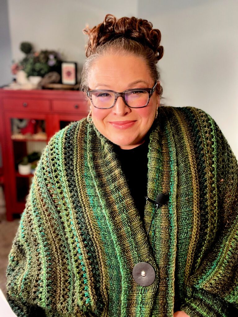 A smiling woman with her hair up in a bun, wearing glasses, showcases the "Know Your Worth Knit Cocoon Cardigan" by Marly Bird. The cardigan features a rich green tone with a subtle lace pattern, accentuated by a large button closure.