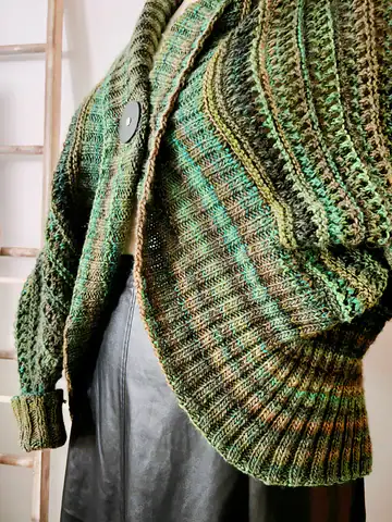 A close-up of a person wearing a textured, multi-shaded green knitted sweater with large buttons over a black leather skirt. The image highlights the cozy fit and intricate knit pattern, perfect for fans of free knitting and crochet patterns. -Marly Bird