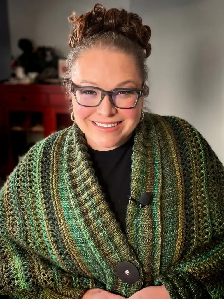 A smiling woman with glasses and curly hair tied up wears a green and brown knitted shawl with large buttons, made from free knitting and crochet patterns. She is indoors, with a blurred cabinet and decorative items in the background. -Marly Bird