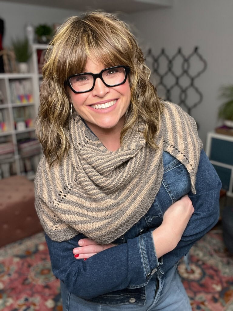 Woman models a striped knit shawl, inspired by Outlander, over a denim jacket in a cozy room with bookshelves and plants.