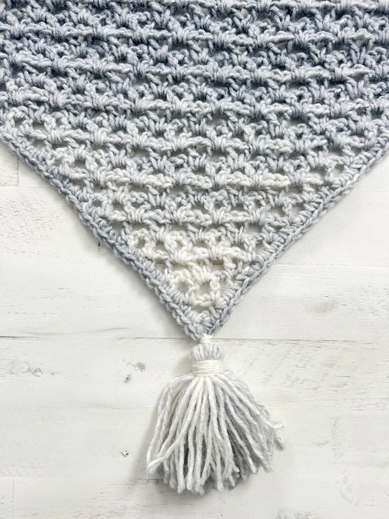 A close-up of a hand-crocheted, triangular shawl with a gradient pattern transitioning from dark gray at the top to white at the bottom. The shawl is adorned with a large white tassel at the bottom point. The shawl is laid out on a light-colored wooden surface. -Marly Bird