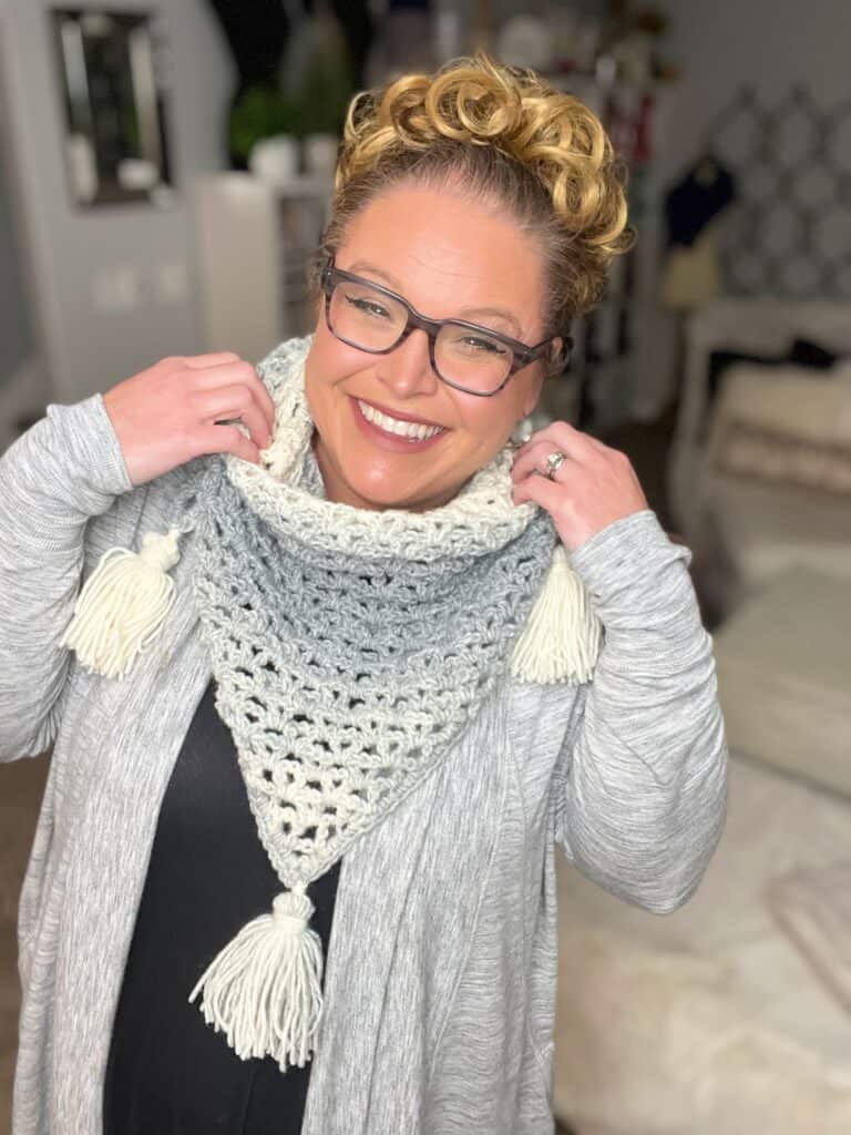 A person with curly hair and glasses smiles while holding up a crocheted triangular scarf with tassels. They are wearing a gray cardigan over a dark shirt. The background appears to be a cozy indoor setting with blurred furniture and decor. -Marly Bird