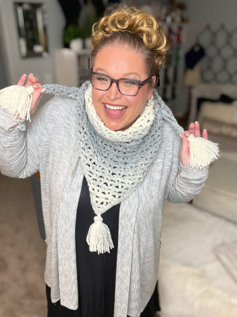 A person with glasses and curly hair is smiling and holding up a light gray and white crocheted scarf with tassels on both ends. They are wearing a gray cardigan over a black top. The background shows a bedroom with a bed and a geometric-patterned wall decor. -Marly Bird