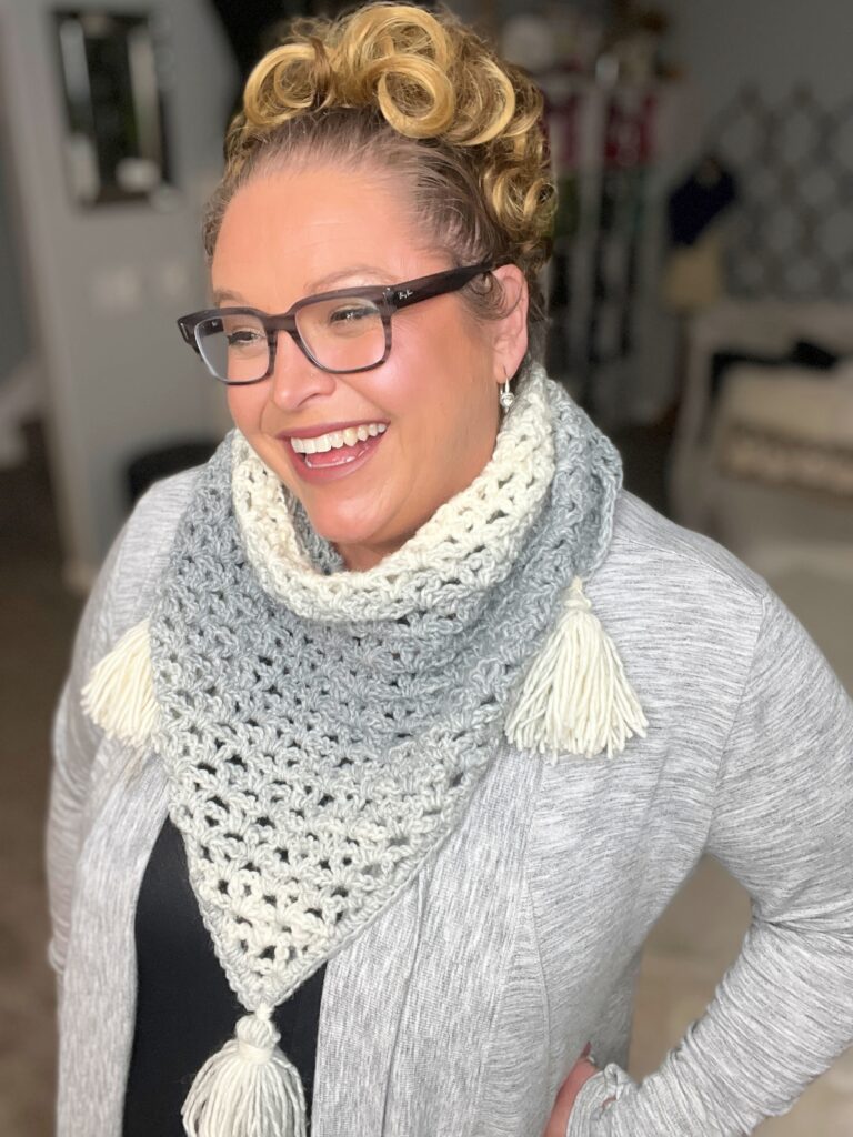 A smiling person with glasses and curly hair is wearing a patterned gray and white scarf with tassels and a light gray cardigan. The background appears to be an indoor setting with blurry household items. -Marly Bird