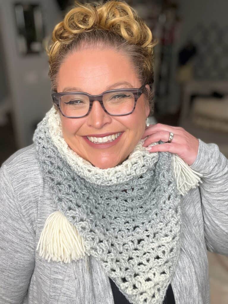 A person is smiling warmly while wearing glasses and a knitted scarf with tassels, featuring shades of grey and white. They are dressed in a light grey sweater, and their hair is styled up. One hand is gently touching the scarf. -Marly Bird