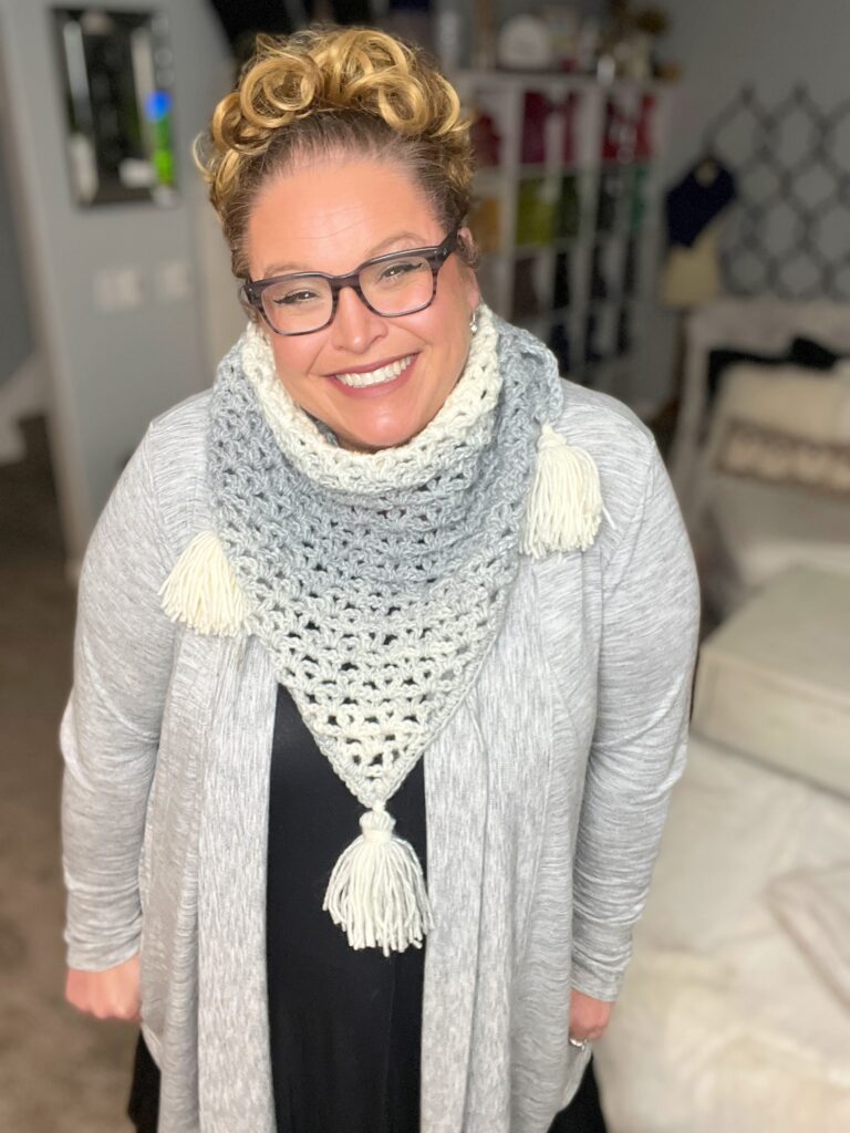 A woman with curly hair wearing glasses smiles warmly at the camera. She is dressed in a gray cardigan and a crocheted scarf with tassels. The background includes a shelf with colorful items and a bed with a patterned headboard. -Marly Bird