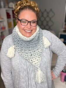 A cheerful woman with glasses and curly hair pulled up is wearing a knitted, gradient grey and white scarf with tassels. She is dressed in a light-grey top and standing in a cozy, well-lit room with shelves in the background. -Marly Bird