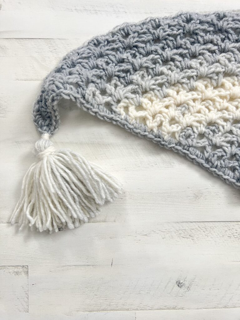 A close-up image of the corner of a crocheted blanket. The blanket features an alternating pattern of grey and off-white yarn and is topped with a white tassel. The crocheted texture and intricate stitch work are visible. The background is a light-colored wooden surface. -Marly Bird